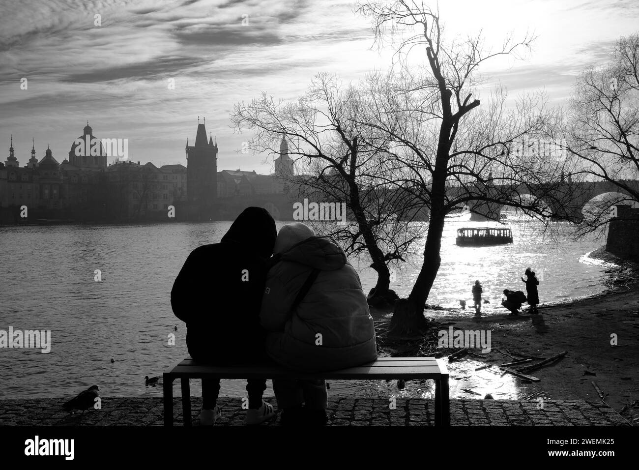 Liebevolles Paar auf einer Bank, die in Prag über den Fluss blickt, während eine Familie am Flussufer spielt Stockfoto