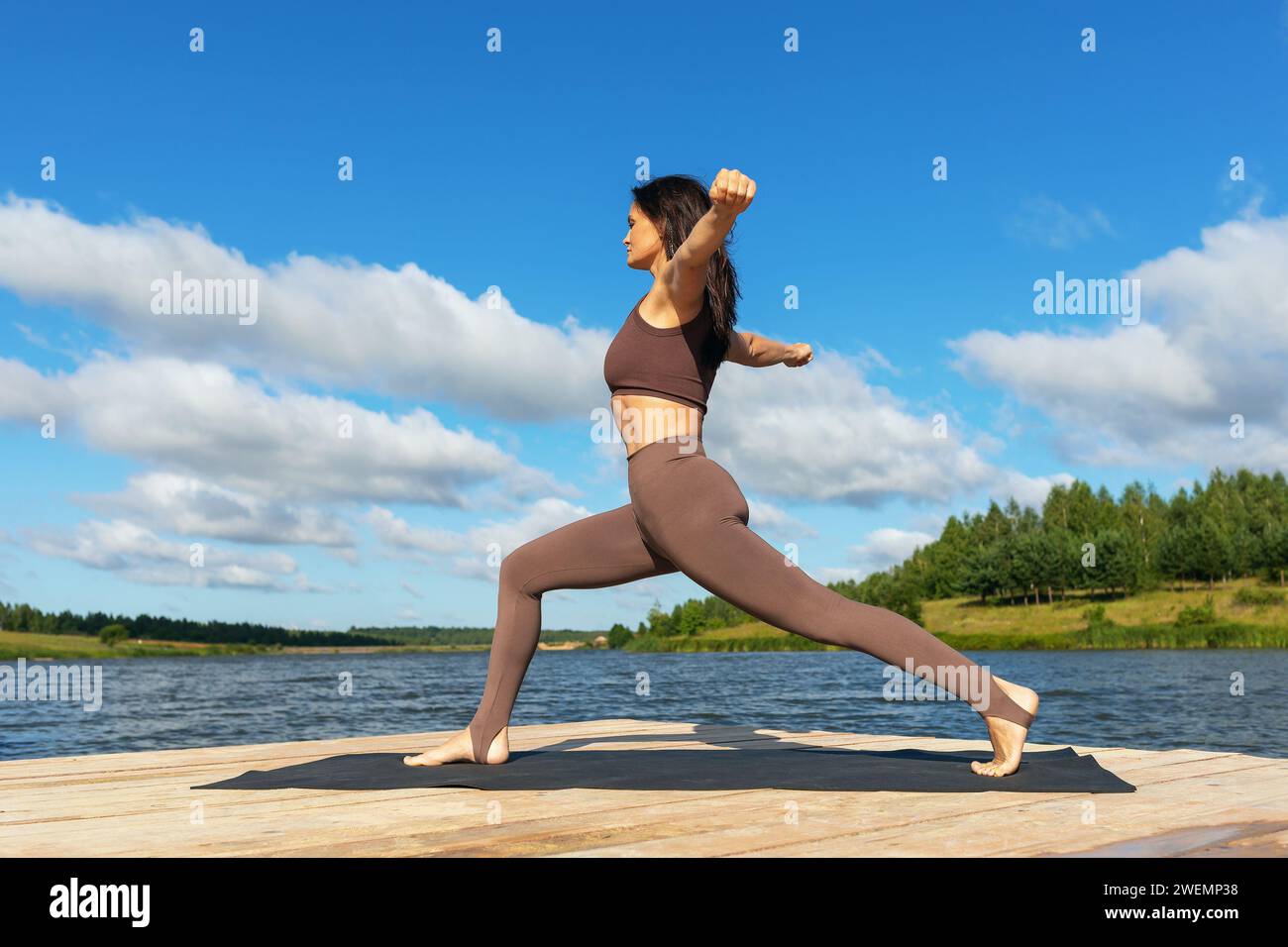 Attraktive Frau, die eine Variation von Virabhadrasana-Übungen, Kriegerstellung, Training in Leggings und einem kurzen Top macht, während sie am Ufer steht Stockfoto