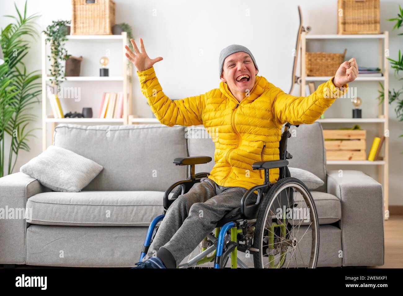 Glücklicher und ausdrucksstarker behinderter Mann im Rollstuhl, der zu Hause die Hände in Freude hebt Stockfoto