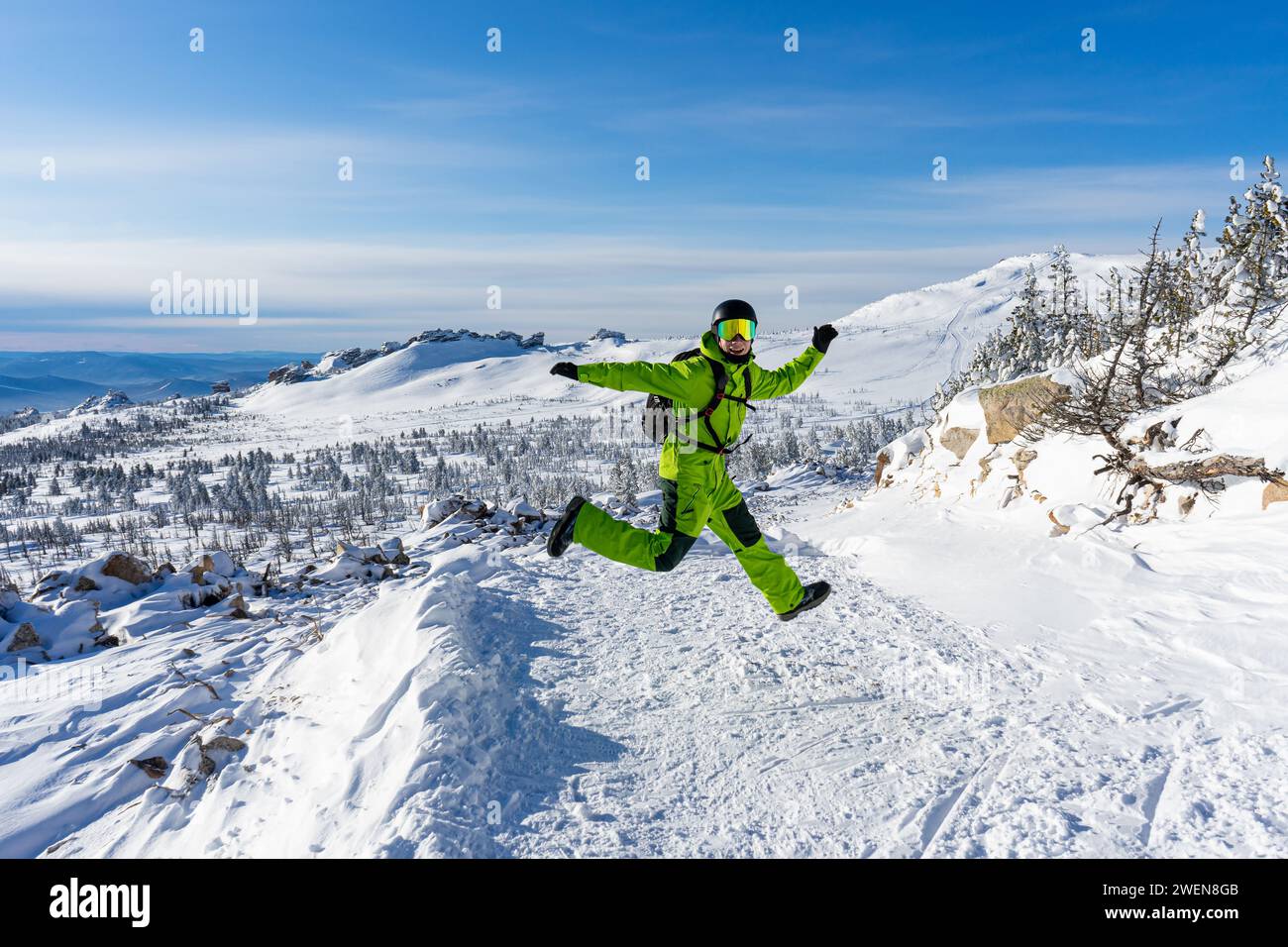 Fröhlicher Mann, der in einem hellgrünen Outfit springt: Warmer Anzug, Schutzbrille, schwarzer Helm, Handschuhe, Rucksack. Schneebedeckte Berge im Skigebiet Sheregesh. Ac Stockfoto