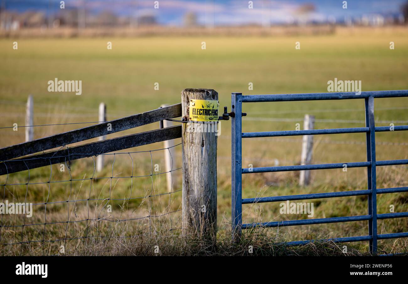 Holzpfosten mit einem elektrischen Zaunschild in landwirtschaftlicher Umgebung, mit einem verzinkten Tor auf der einen Seite und einem Zaun auf der anderen Seite Stockfoto