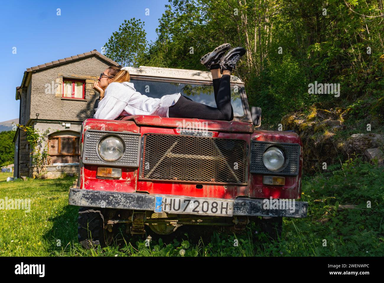 Eine Frau schaut nachdenklich in den Himmel über der Motorhaube eines alten roten Geländewagens, seitlich Stockfoto
