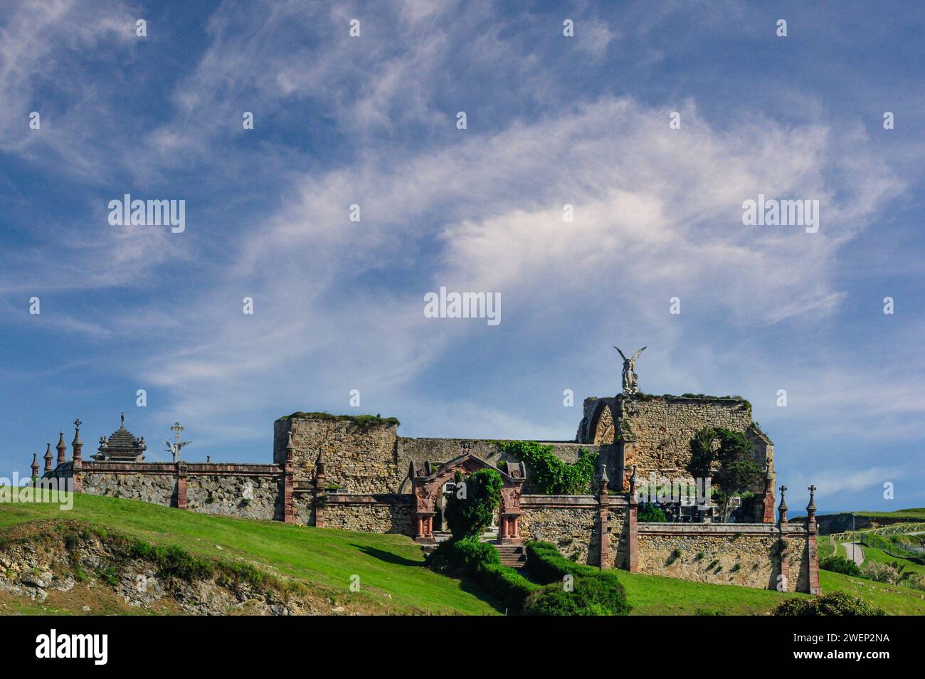 Auf den alten Steinmauern des historischen Friedhofs in Comillas steht unter einem dynamischen Himmel eine Statue eines befehlenden Engels Stockfoto