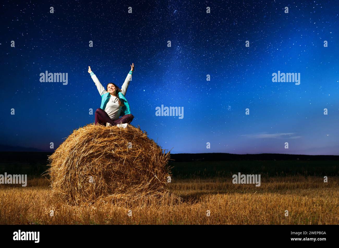 Das junge glückliche Mädchen mit erhobenen Armen sitzt auf einem runden Heuhaufen auf einem Feld vor dem Hintergrund des Sternenhimmels. Nächtliche Landschaft des Sternenhimmels mit Stockfoto