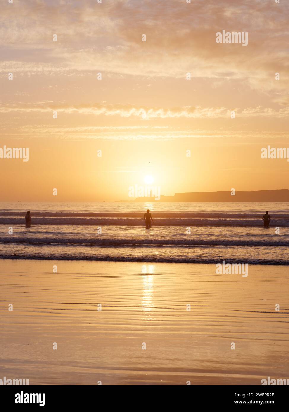 Menschen, die im Meer baden, mit Insel dahinter bei Sonnenuntergang in Essaouira, Marokko, 26. Januar 2024 Stockfoto