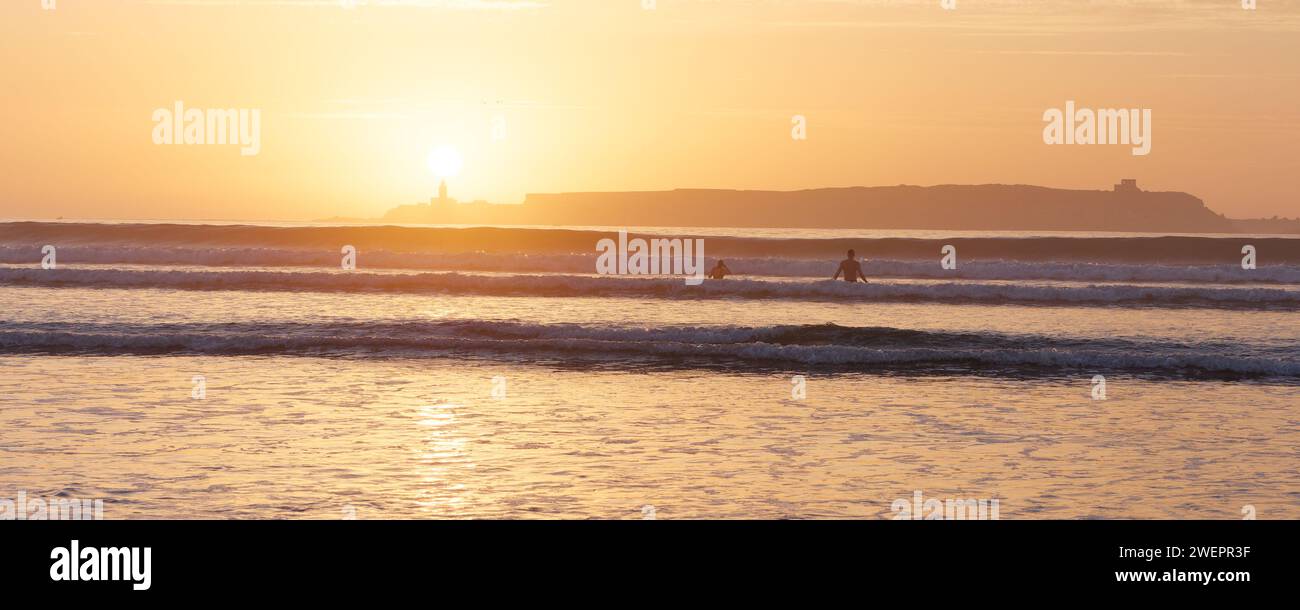 Menschen, die im Meer baden, mit Insel dahinter bei Sonnenuntergang in Essaouira, Marokko, 26. Januar 2024 Stockfoto
