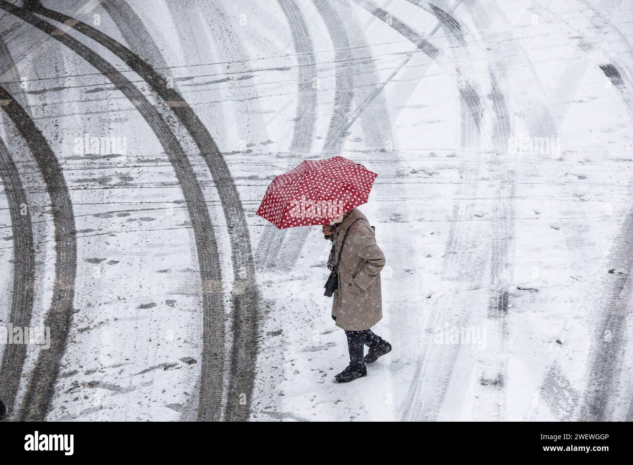 Frau mit einem rot-weiß gepunkteten Schirm überquert eine schneebedeckte Straße im Stadtzentrum, Schnee, Winter, Köln, Deutschland. Januar: 2024 Frau mit Stockfoto