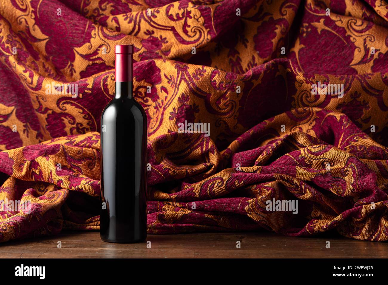 Eine Flasche Rotwein auf einem alten Holzteller. Im Hintergrund Retro-Wandteppich mit dunkelrotem und goldenem Blumenschmuck. Stockfoto