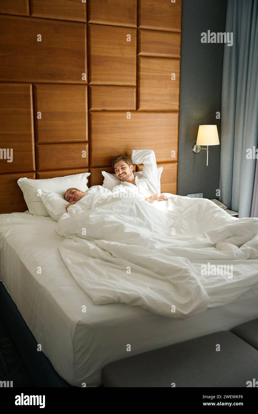 Die blonde Frau und ihr Freund wachen auf einem großen Bett auf, sie mieten ein gemütliches Hotelzimmer Stockfoto