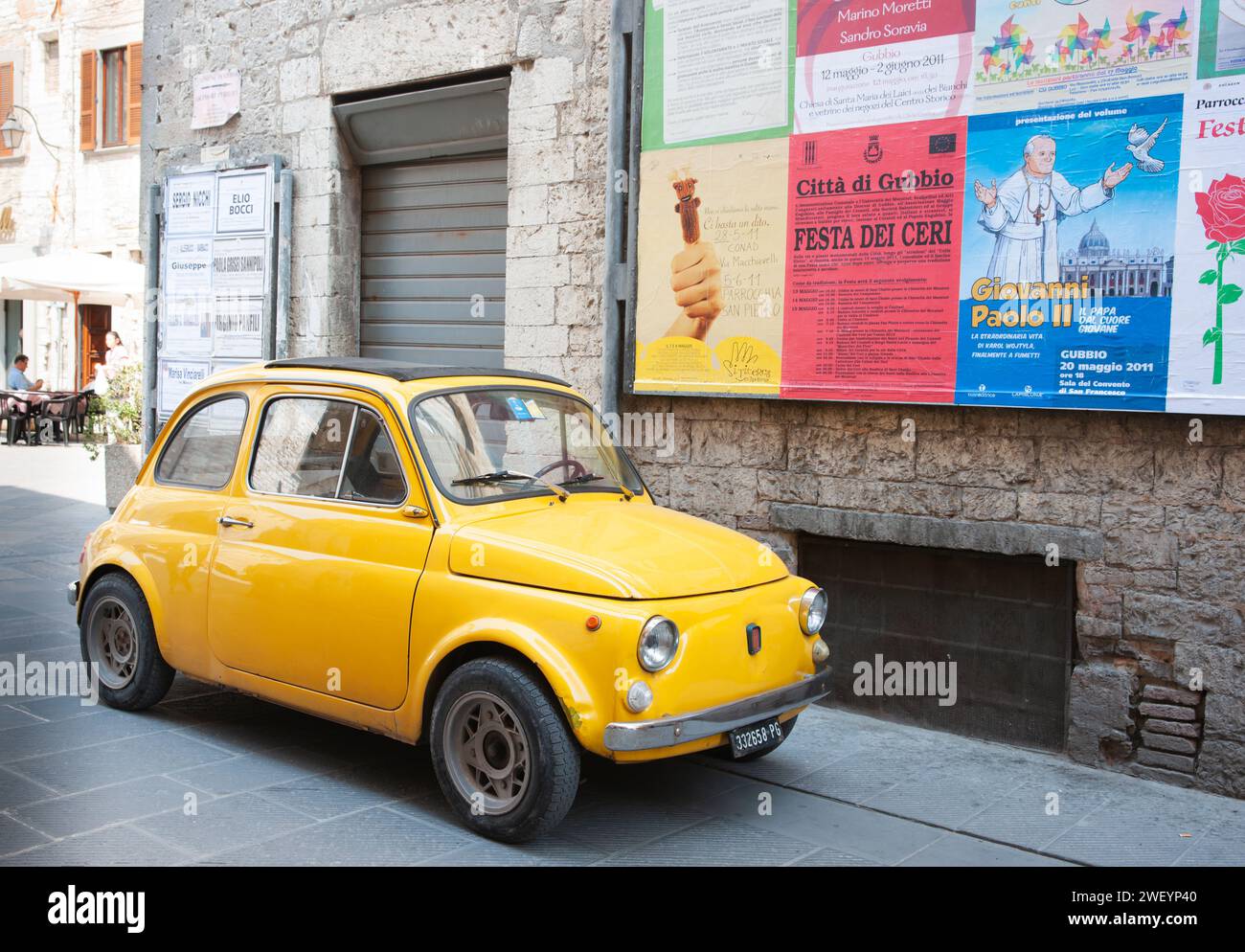 Gubbio Italien - 13. Mai 2011; alter gelber kleiner italienischer Oldtimer parkt in der Altstadtstraße unter einer Wand von Plakaten in italienischer Sprache. Stockfoto