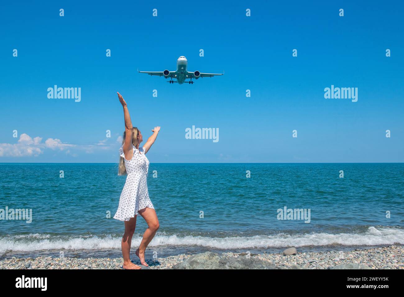 Parallele Reisen: Mädchen in weißem Kleid auf Steinen, Flugzeug überquert das Blaue Meer Stockfoto