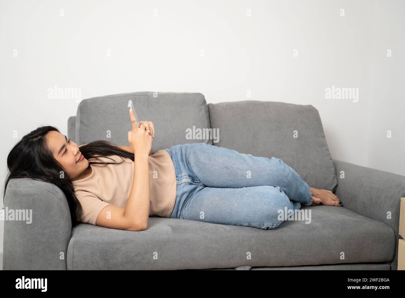 Asiatische junge Frau mit Handy, die auf dem Sofa liegt. Stockfoto