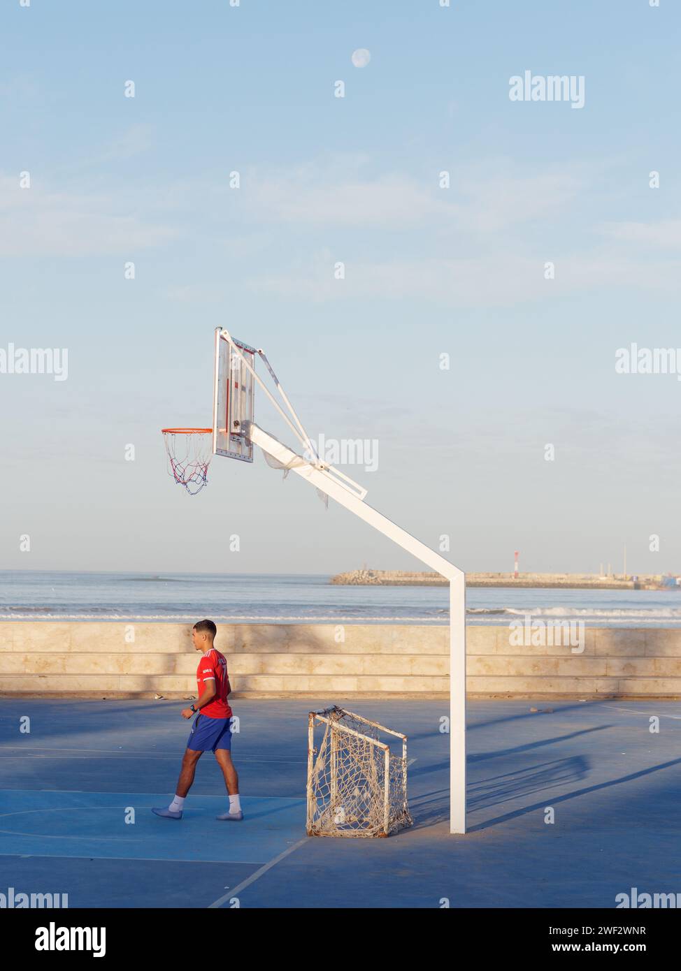 Junger Torhüter mit provisorischem Tor auf einem blauen Basketballfeld mit dem Atlantik dahinter und dem Mond darüber in Essaouira, Marokko, 28. Januar 2024 Stockfoto