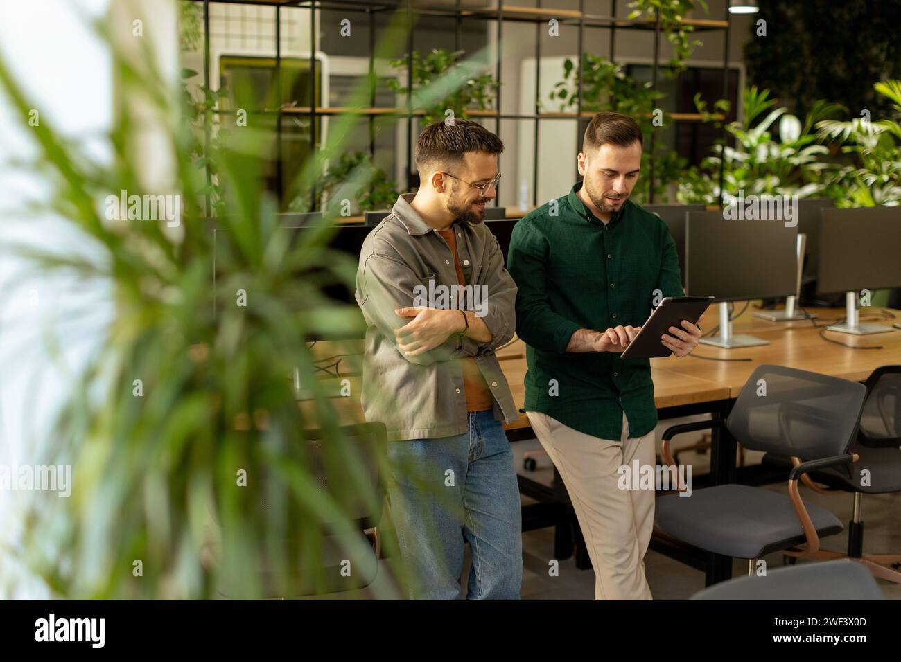 Zwei Profis führen ein fokussiertes Gespräch, während sie ein digitales Tablet in einem modernen Bürobereich mit rustikalem, freiliegendem Mauerwerk halten Stockfoto