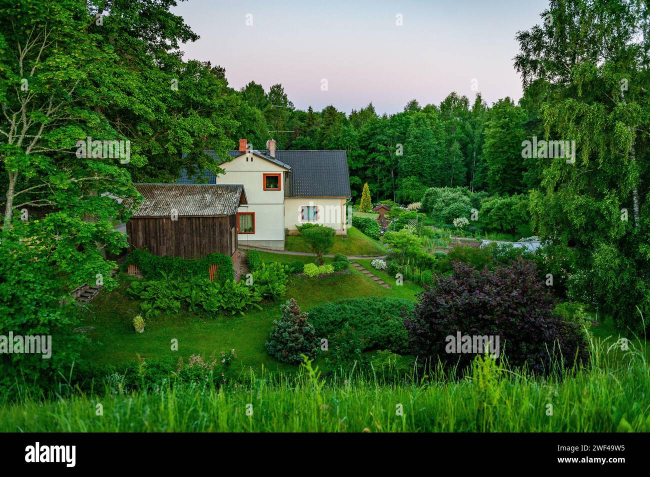Schöne Hinterhoflandschaft mit gepflegtem Rasen, Blumenbeeten, Holzscheune und Haus bei Sonnenaufgang. Smiltene, Lettland. Stockfoto