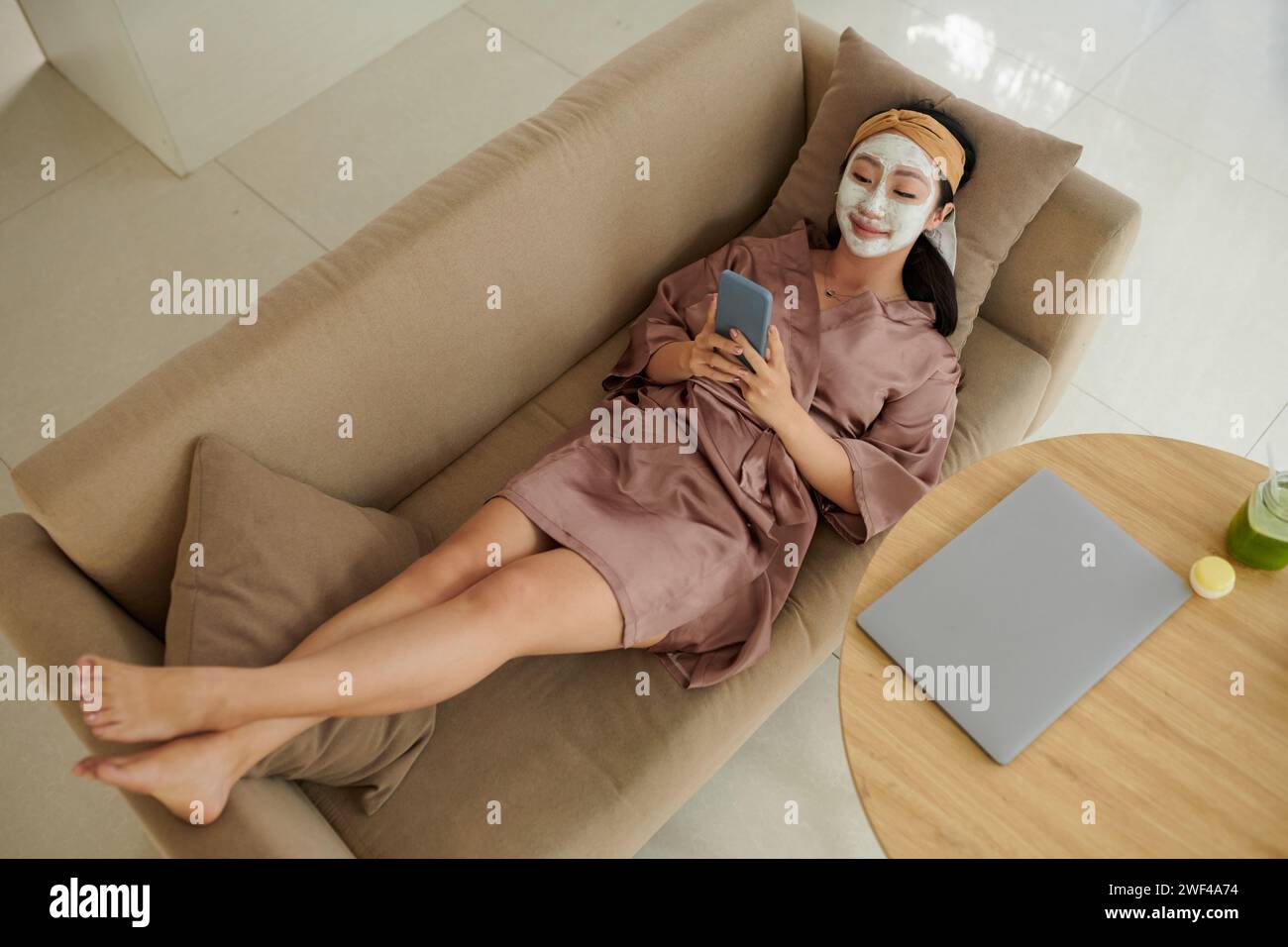 Glückliche junge Frau im Seidenmantel, die sich auf der Couch mit Gesichtsmaske auf und einem Freund eine SMS schickt Stockfoto
