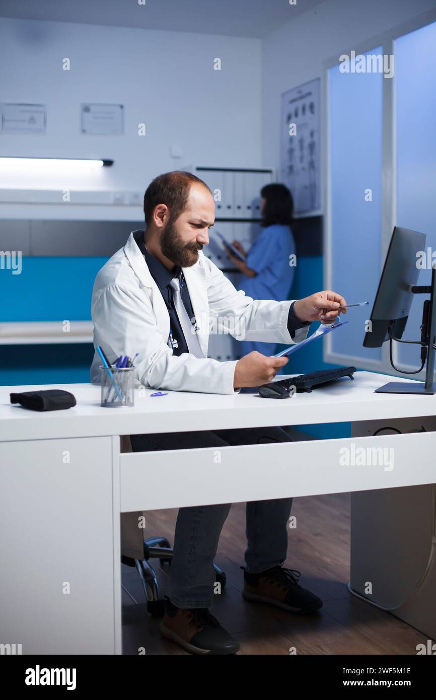 Detailliertes Bild eines männlichen Arztes am Klinikpult, der seine Notizen analysiert. Kaukasischer Mann, der Desktop-Computer und Zwischenablage verwendet, während er sich auf die medizinischen Konsultationen des Patienten vorbereitet. Stockfoto