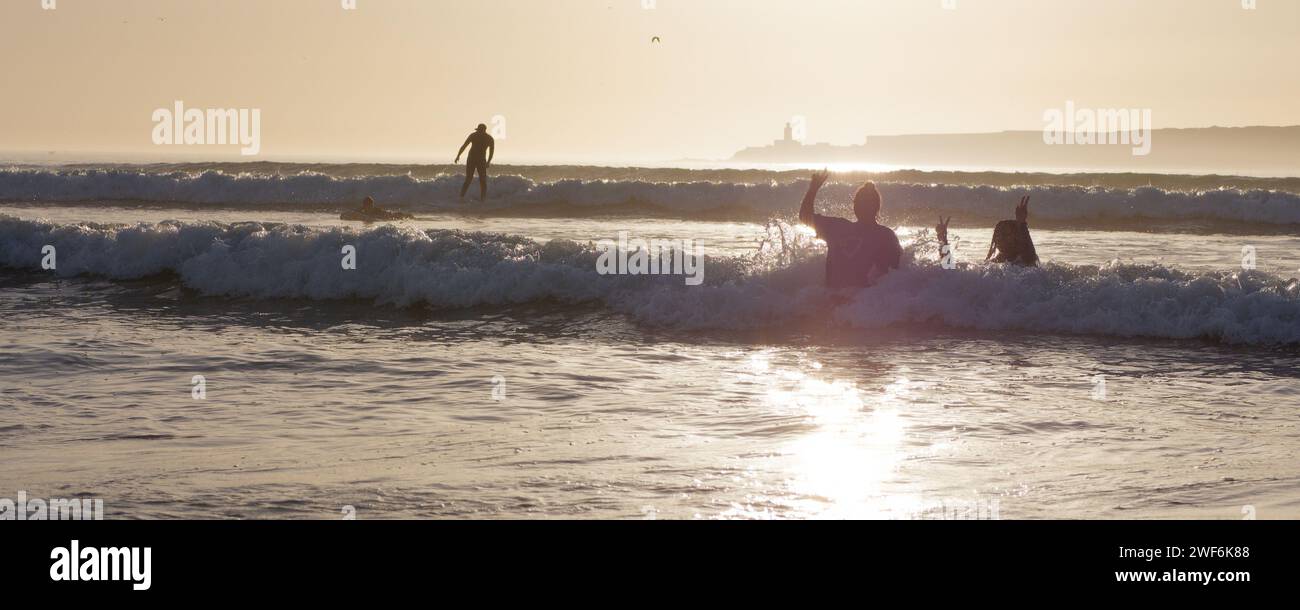 Surfer in Silhouette reitet eine Welle bei Sonnenuntergang mit Menschen im Meer im Vordergrund mit einer Insel dahinter in Essaouira, Marokko, 28. Januar 2024 Stockfoto