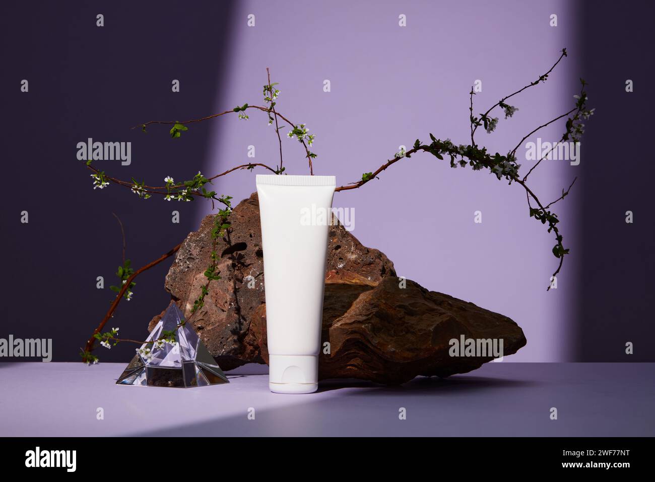 Vorderansicht des weißen Kunststoffröhrchens mit Gesichtscreme oder Gesichtsreiniger auf violettem Hintergrund. Brauner Stein mit weißer Blume als Highlight. Stockfoto