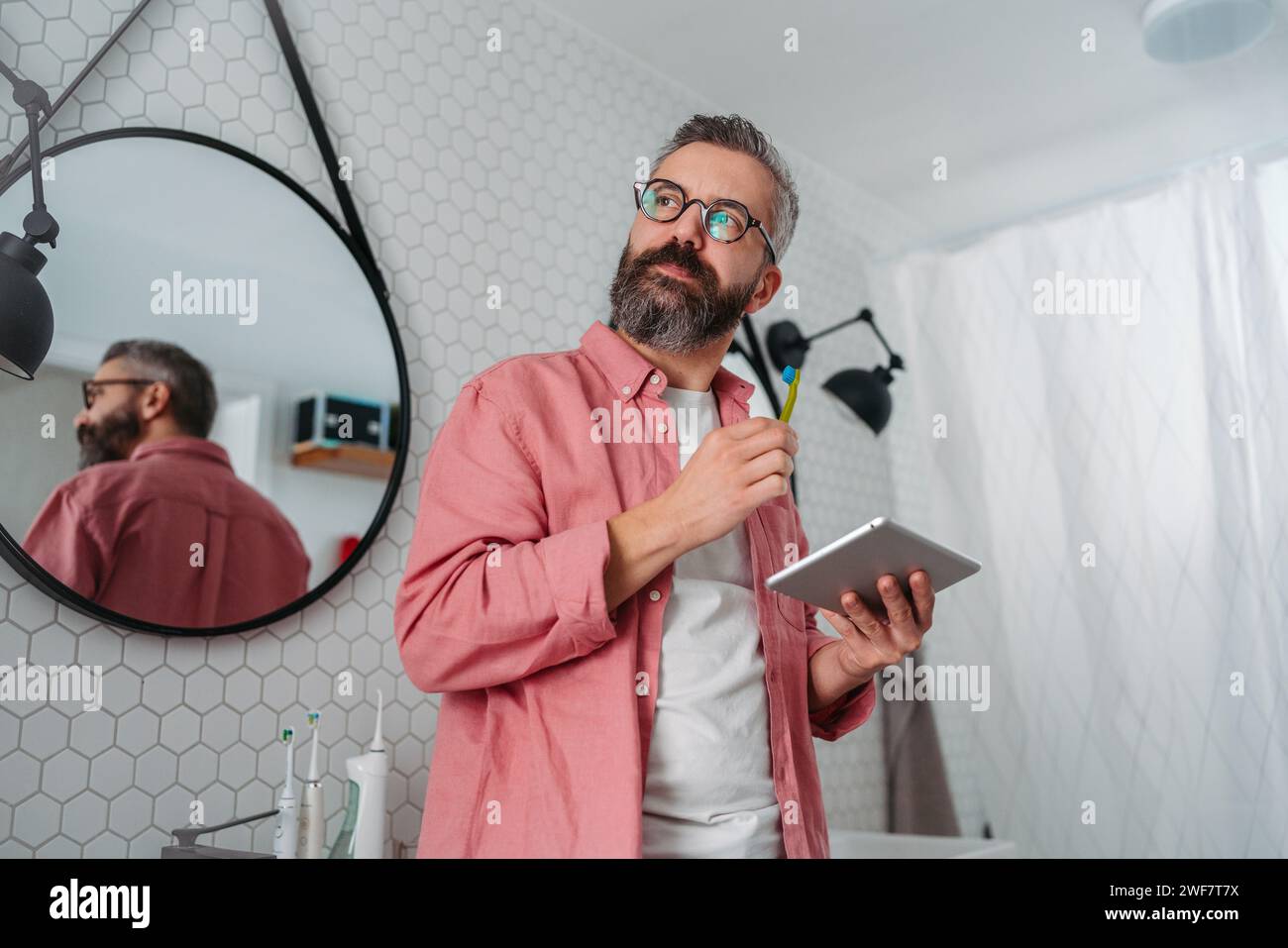 Ein Mann, der online einkaufen und sich im Badezimmer die Zähne putzt. Tablette halten, scrollen und kaufen, Zahnbürste im Mund. Stockfoto