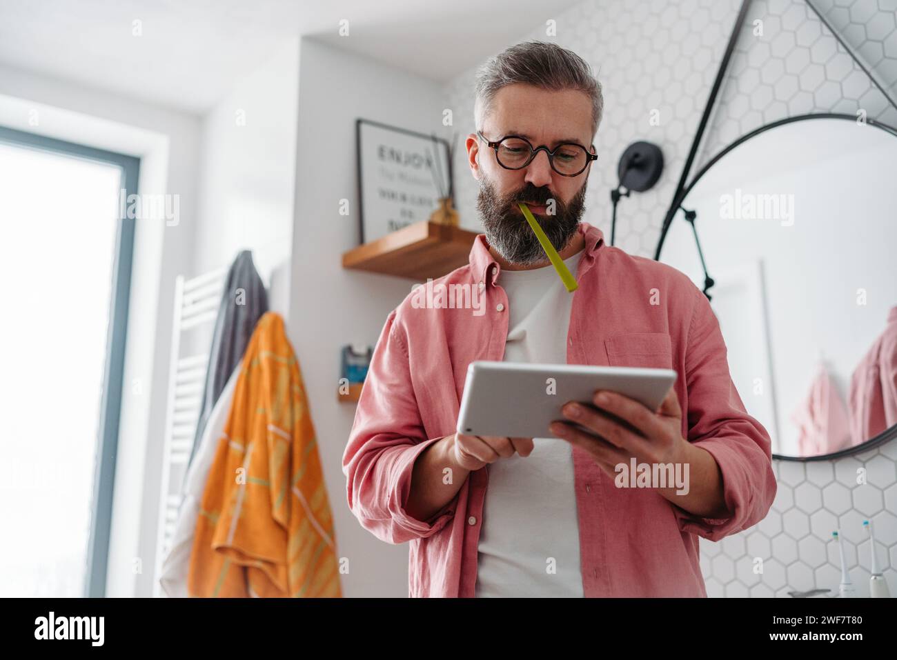 Ein Mann, der online einkaufen und sich im Badezimmer die Zähne putzt. Tablette halten, scrollen und kaufen, Zahnbürste im Mund. Stockfoto