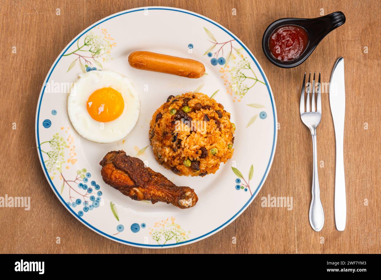 Amerikanischer gebratener Reis mit gebratenem Trommelstock, Spiegelei und Wurst auf weißer Keramikplatte mit Ketchup in schwarzer Tasse, Metallgabel und Messer auf Holztisch Stockfoto