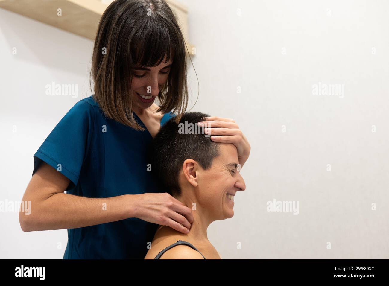 Frau in blauen Peelings gibt einer lächelnden Empfängerin eine entspannende Kopfmassage Stockfoto
