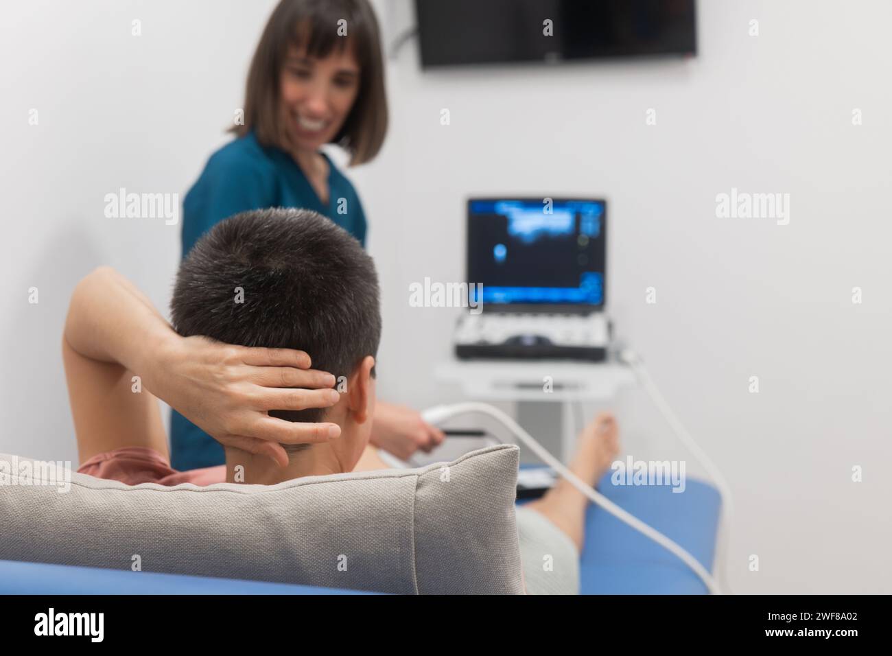 Ein männlicher Patient unterzieht sich einer Ultraschalltherapie mit einer lächelnden Physiotherapeutin in einer klinischen Umgebung Stockfoto