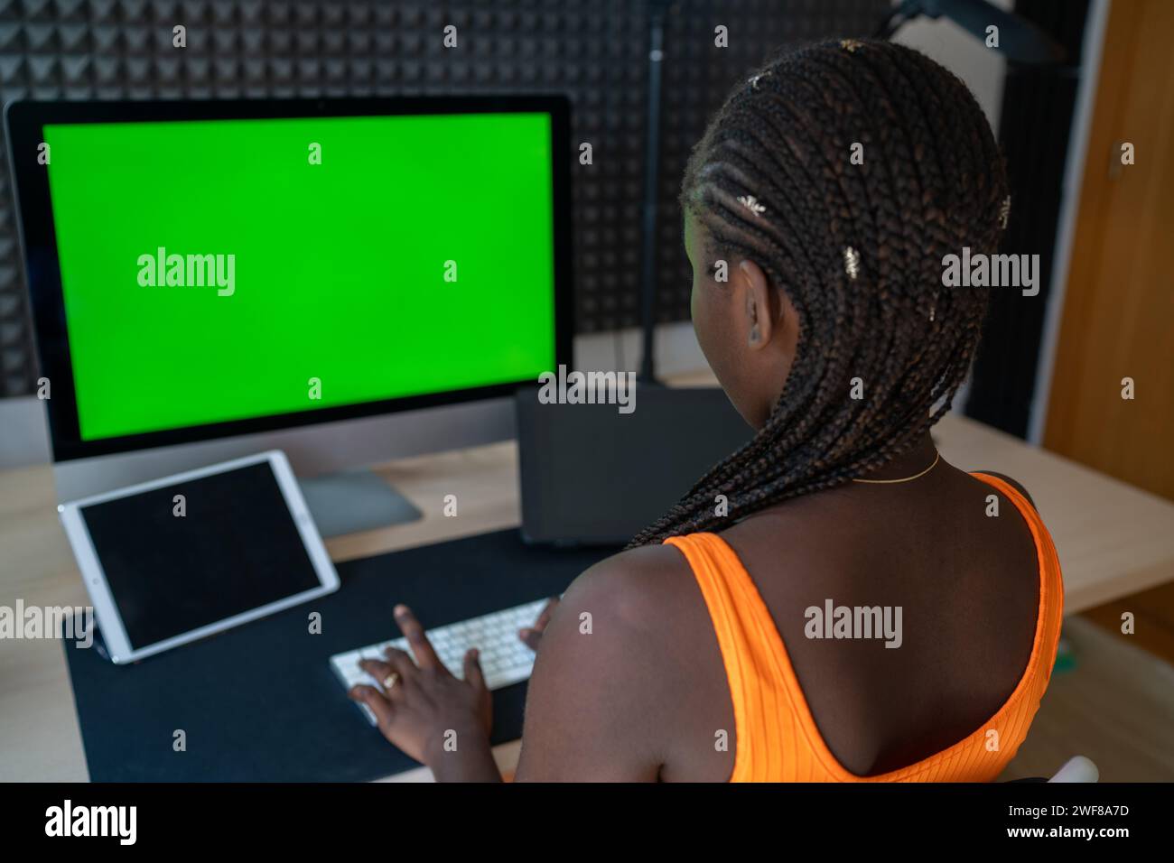Rückansicht einer jungen Dame in orangefarbenem ärmellosem Kleid, die Informationen mit der Tastatur auf dem Schreibtisch eingibt, während sie während der vac-Funktion ferngesteuert vom modernen Arbeitsplatz aus arbeitet Stockfoto