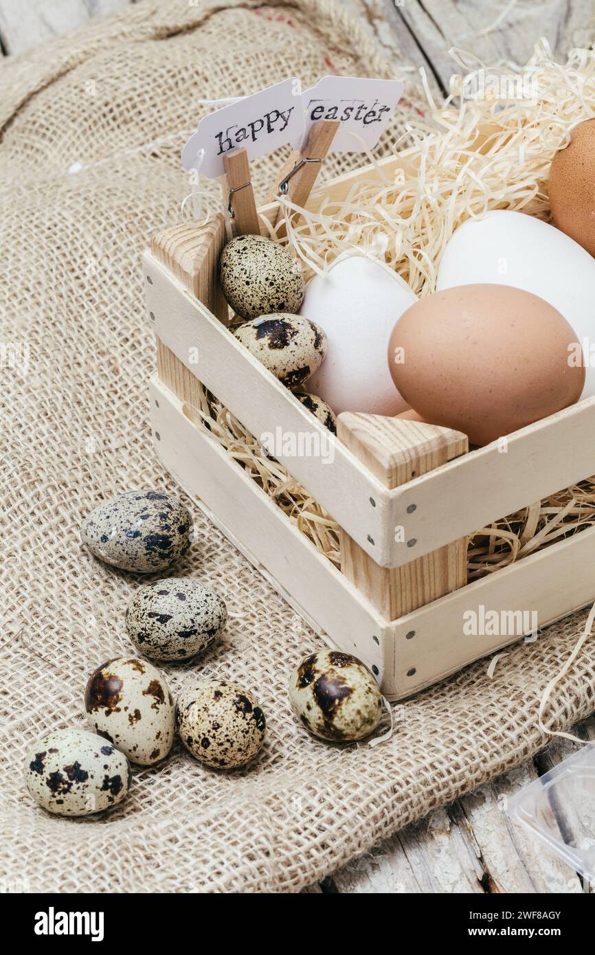 Ein Holzkorb gefüllt mit verschiedenen Eiern und ein Schild mit der Aufschrift „Happy Easter“ auf rustikalem Sackleinen. Stockfoto