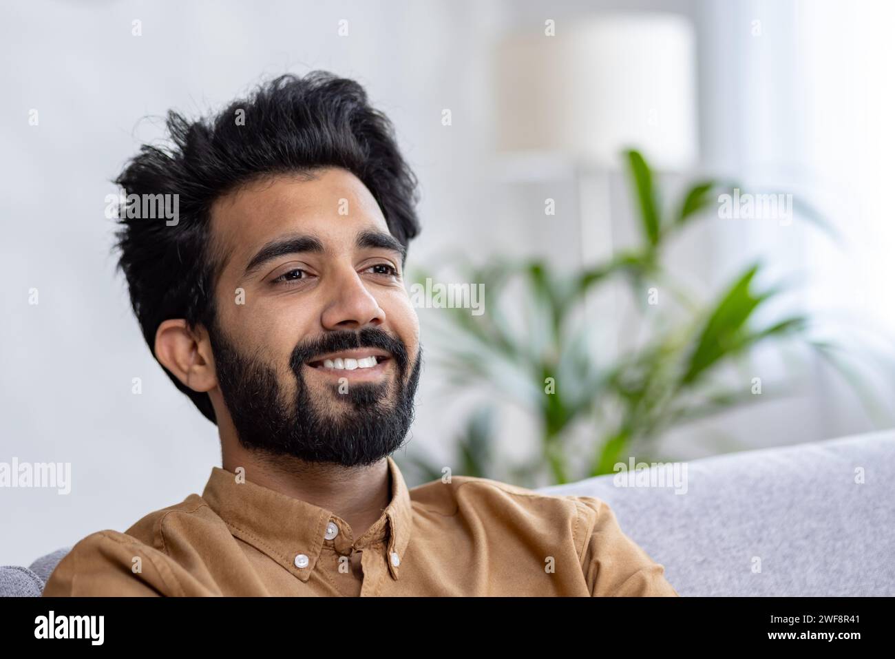 Ein zufriedener junger Indianer sitzt zu Hause auf dem Sofa und ruht aus. Ein lächelnder Mann schaut zur Seite, Nahaufnahme. Stockfoto