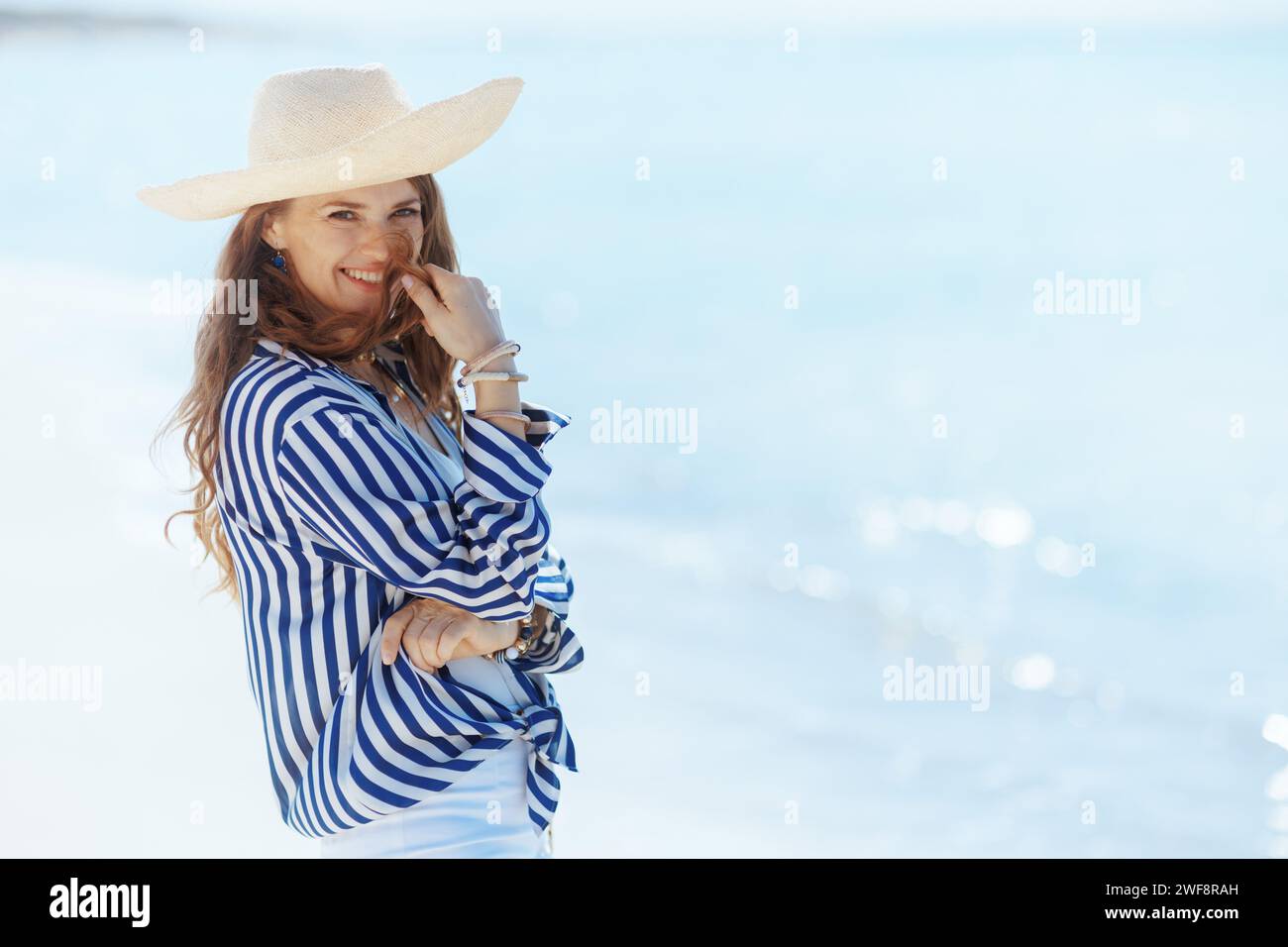 Lächelnde, moderne Frau mittleren Alters an der Küste mit Strohhut, die Spaß hat. Stockfoto