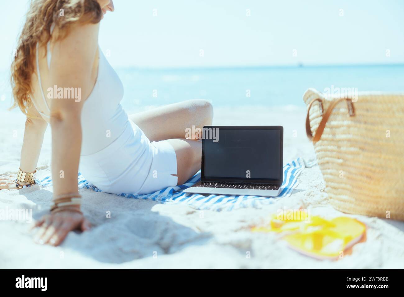 Entspannte, stilvolle Frau am Meer mit Strohtasche, leerem Laptop-Bildschirm und gestreiftem Handtuch. Stockfoto