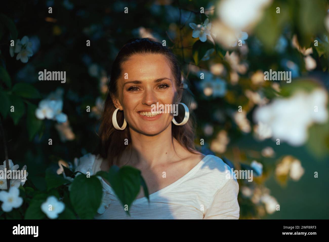 Sommerzeit. Porträt einer lächelnden eleganten 40-jährigen Frau in weißem Hemd in der Nähe des blühenden Baumes. Stockfoto