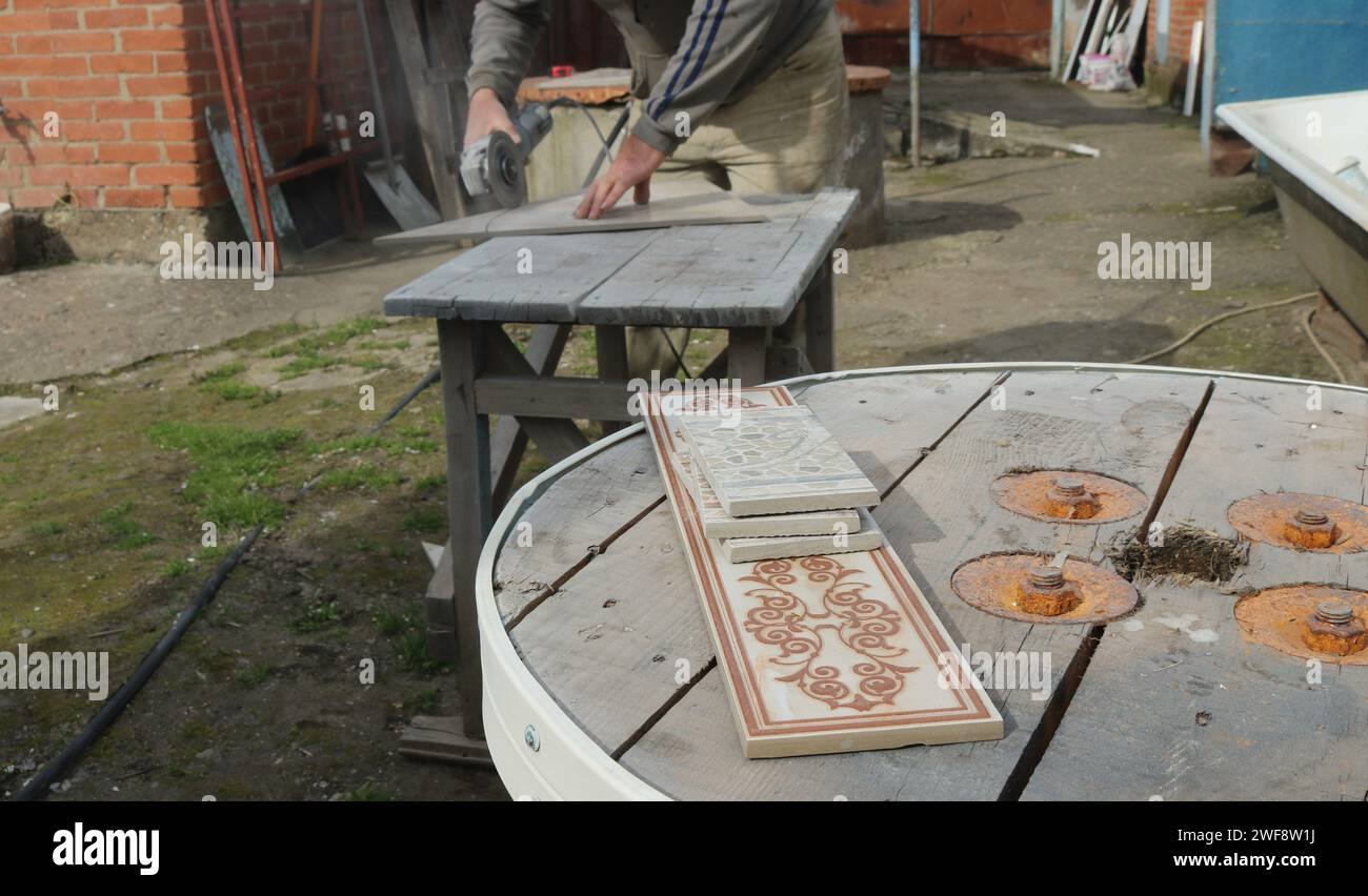 Dekorative Fliesen, die auf einer Holzfläche auf der Straße vor einem verschwommenen Hintergrund eines Arbeitsprozesses liegen, bei dem ein Handwerker Fliesen verarbeitet Stockfoto