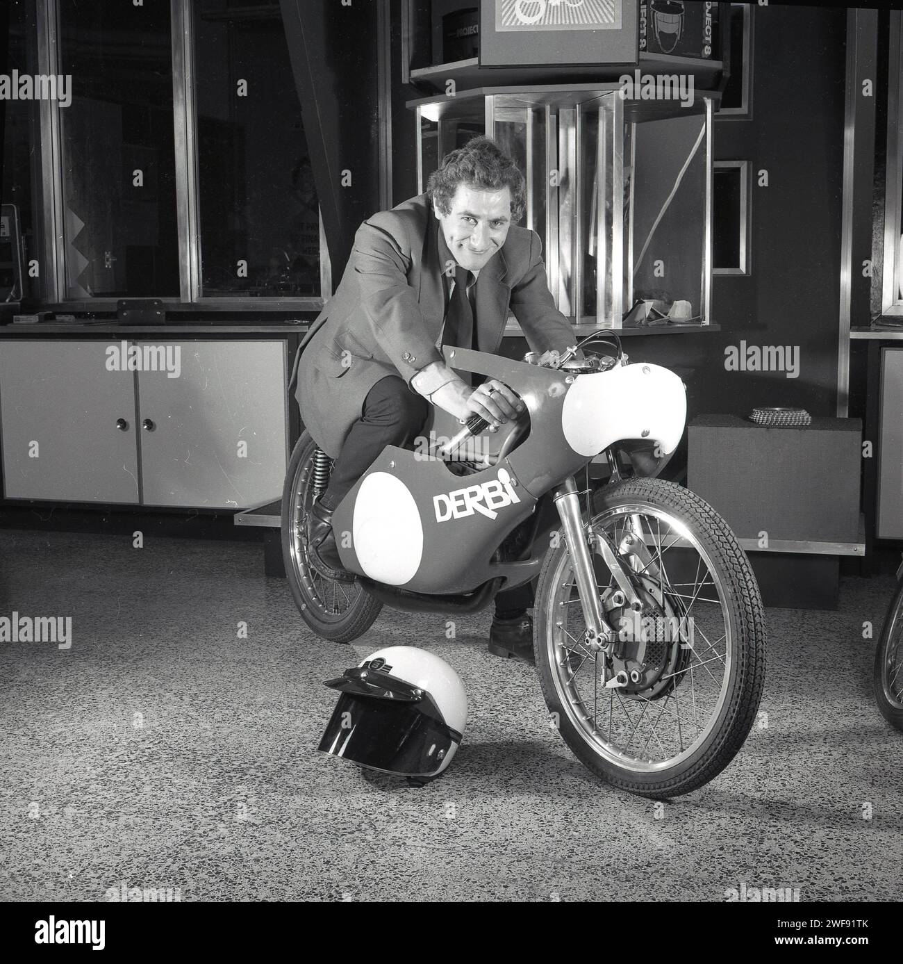 1974, historisch, ein passender Automobilverkäufer, der auf einem DERBI 500GP Motorrad in einem Autohaus sitzt, England, Großbritannien. Ursprünglich als Fahrradreparaturwerkstatt gegründet - DERBI leitet sich von dem Begriff 'Derivats de Biccletes' ab - in einem Dorf in der Nähe von Barcelona in den 1920er Jahren begann das Unternehmen mit der Herstellung von Mopeds in den 1940er Jahren 1950 wurde er als Nacional Motor SA bekannt und brachte sein echtes Motorrad, den DERBI 250, vor, das später in den späten 60er und frühen 70er Jahren in der 50er-Klasse große Erfolge beim Grand Prix-Motorradrennen erlebte. Stockfoto
