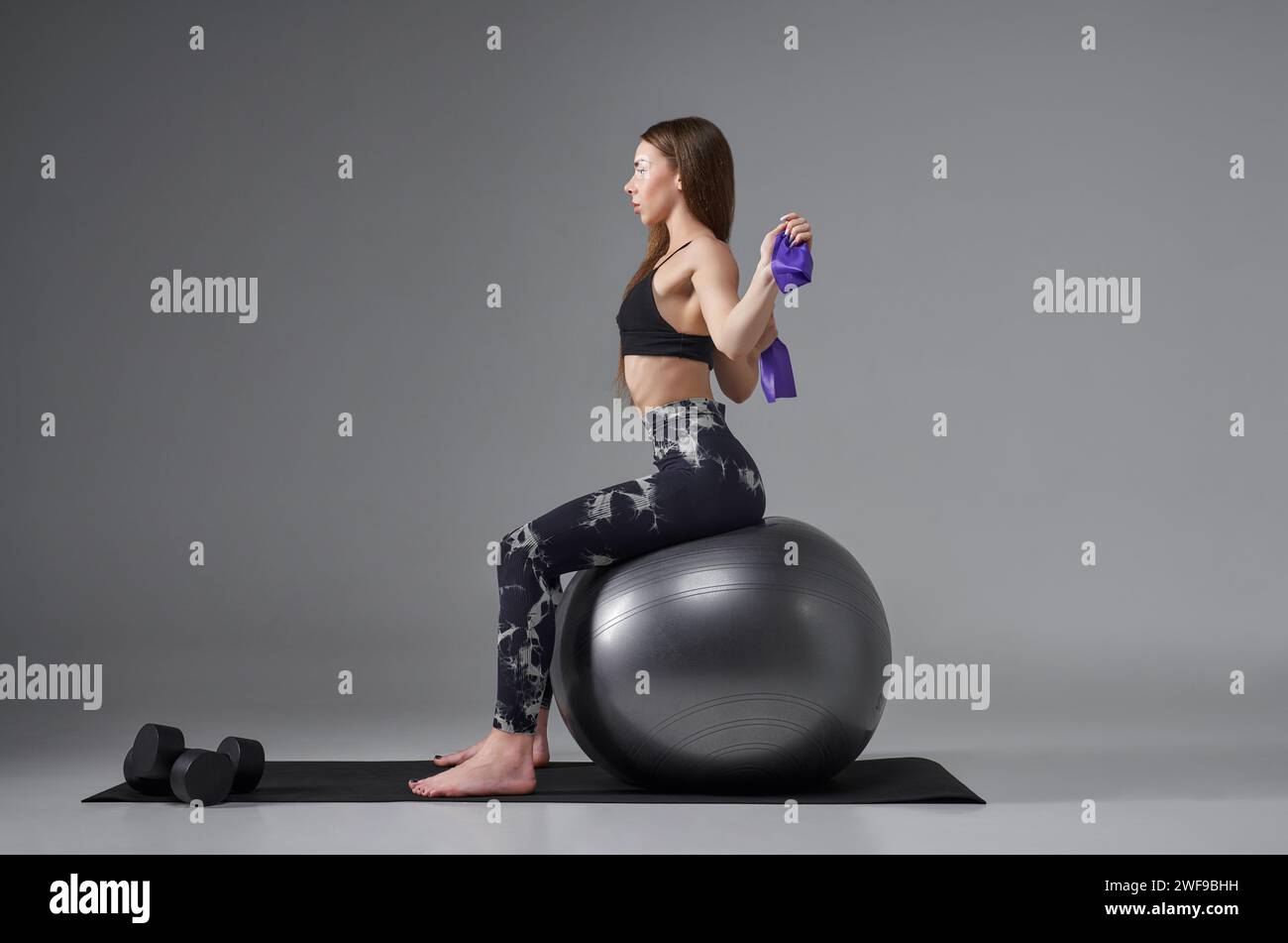 Entschlossene junge Frau in Aktivkleidung mit Gummiband, während sie die Arme hinter dem Rücken spannt. Seitenansicht der weiblichen Athletin, die auf einem Fitnessball trainiert, IS Stockfoto