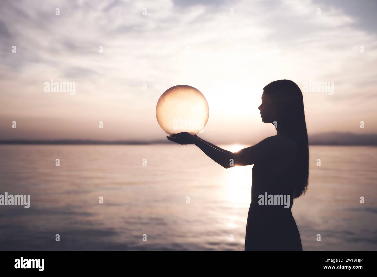 Die Frau hält mit ihren Händen eine surreale Kugel, die von der Sonne beleuchtet wird, die Energie freisetzt, abstraktes Konzept Stockfoto