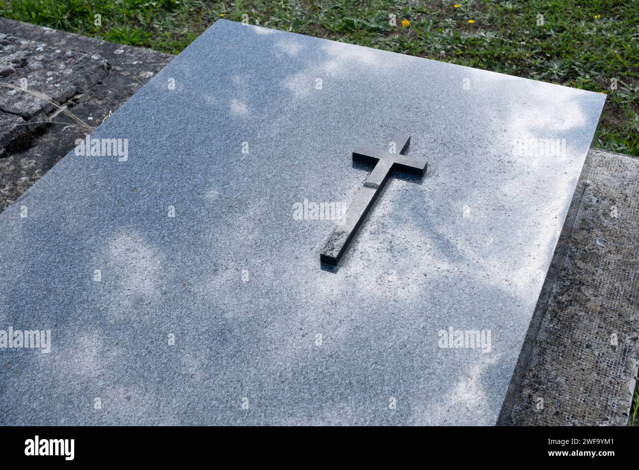 Einfaches dunkles Kreuz auf einem glänzenden Marmorgrabstein auf einem Friedhof. Wolken spiegeln sich in der Oberfläche der Platte Stockfoto