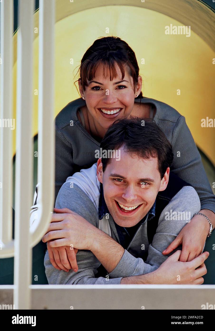 Ein fröhlicher Mann und eine Frau umarmen sich, während sie durch einen Spielplatztunnel schauen, beide tragen legere Kleidung und teilen einen Moment des Glücks Stockfoto