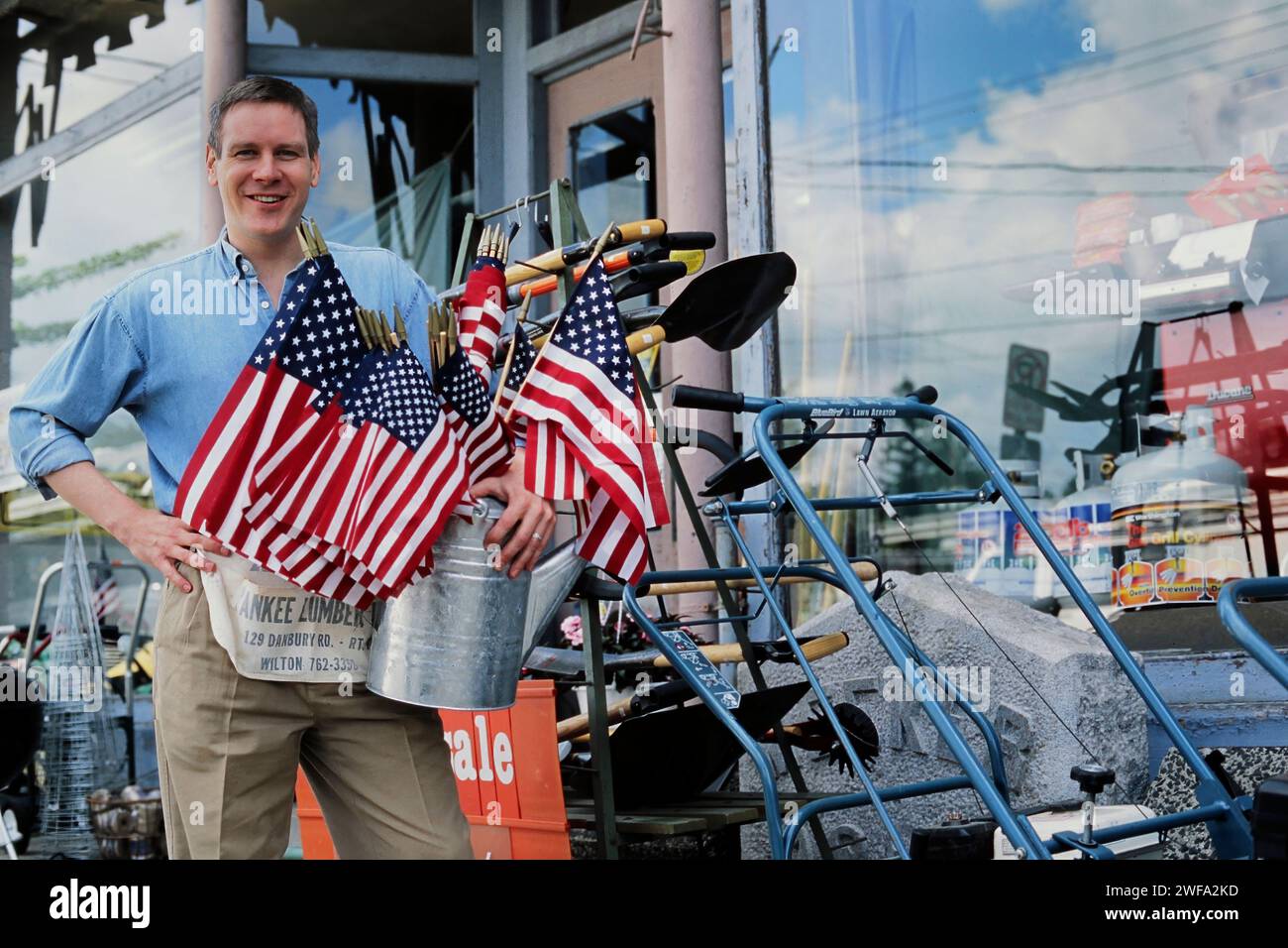 Ein Porträt des stolzen Besitzers eines Kleinbaugeschäfts, der am Eingang seines Geschäfts steht und einen Eimer amerikanischer Flaggen hält Stockfoto