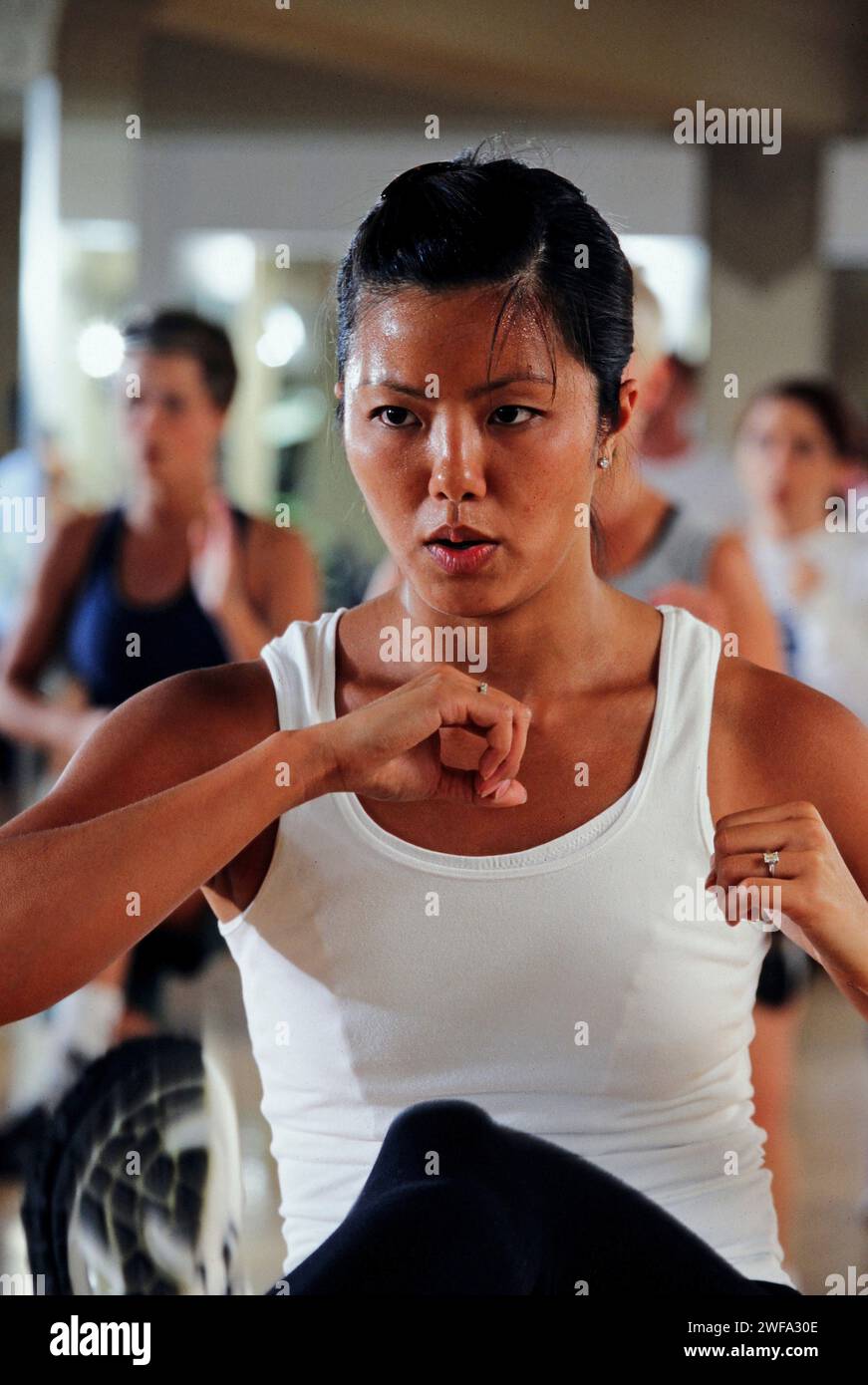 Eine entschlossene Frau wird in einer Notwehr-Haltung während eines Kampfsporttrainings in einem Fitnessstudio mit anderen Teilnehmern im Hintergrund dargestellt. Stockfoto
