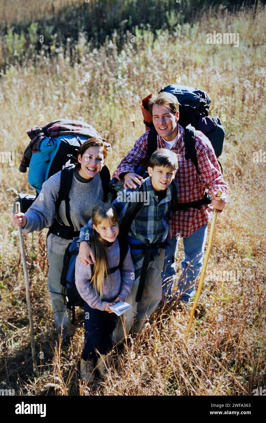 Ein Porträt einer glücklichen Familie mit zwei Kindern und Rucksäcken lächelt die Kamera an, während Sie an einem sonnigen Herbsttag auf einem grasbewachsenen Feld wandern. Stockfoto