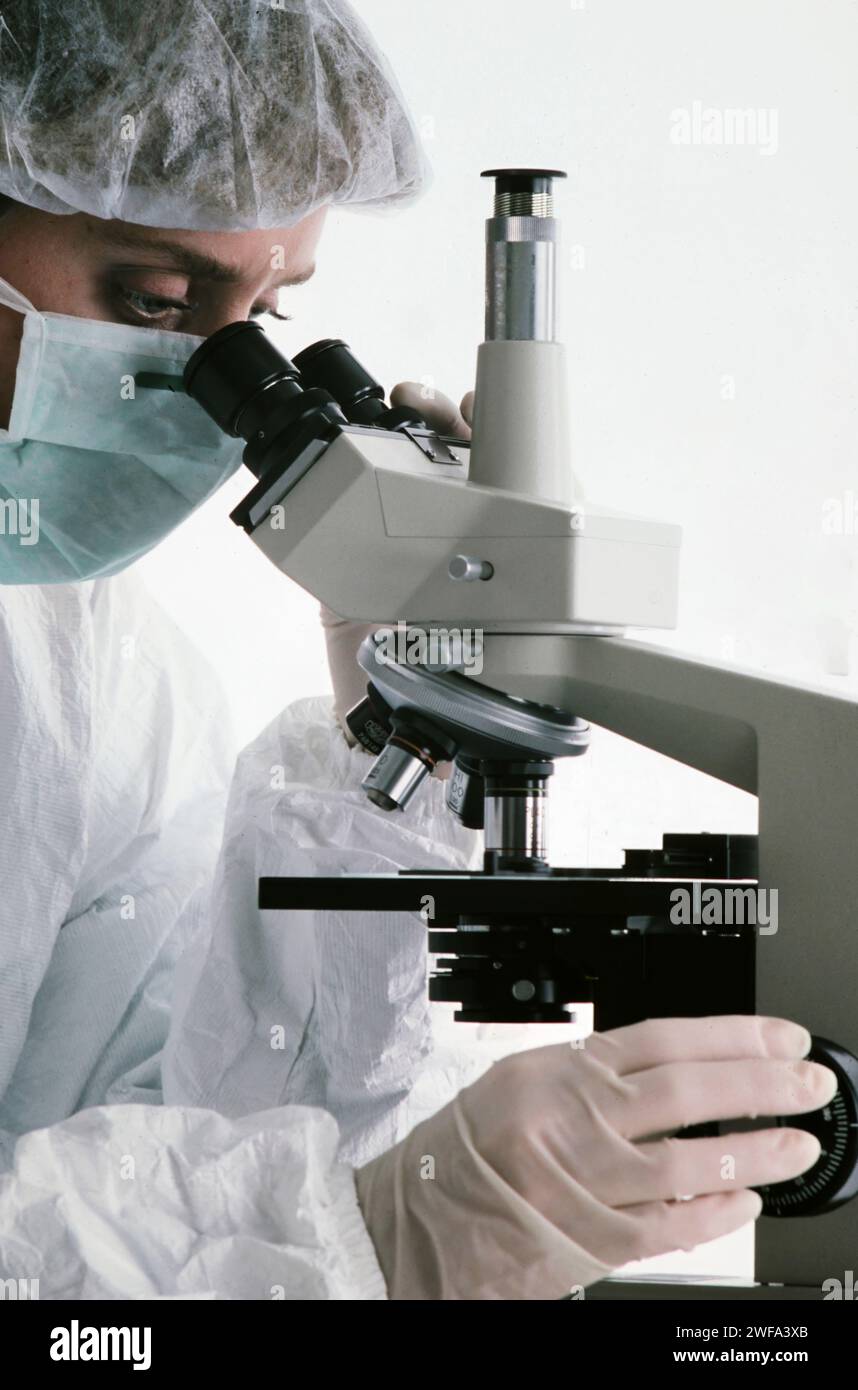 Ein fokussierter Wissenschaftler, der in einen Labormantel und eine Schutzmaske gekleidet ist, untersucht eine Probe sorgfältig durch die Linse eines modernen Mikroskops in einer sauberen Arbeit Stockfoto