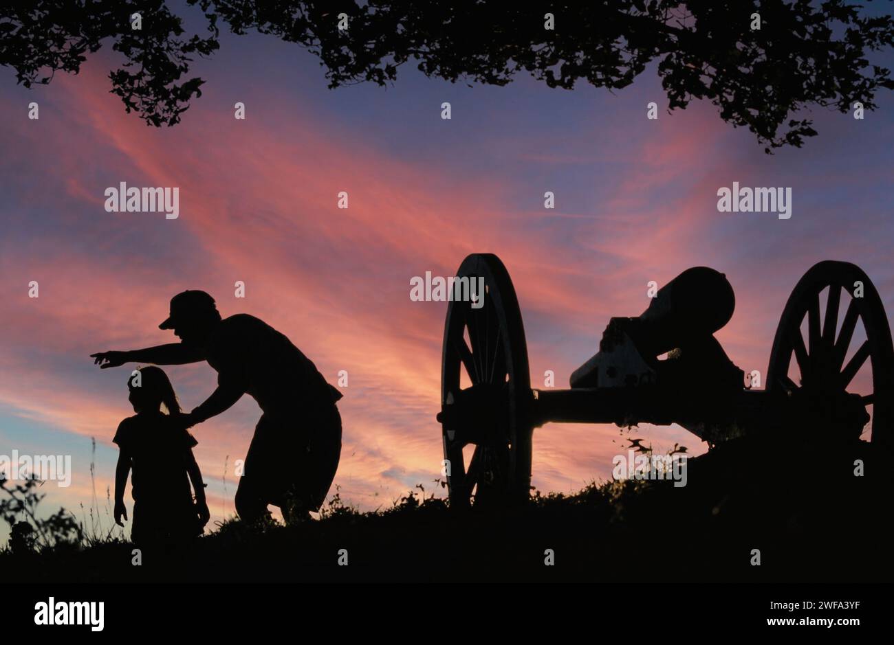 Ein Vater und eine Tochter, die in Silhouette vor einem lebendigen Sonnenuntergang gefangen wurden, während sie die historische Bürgerkriegsartillerie auf einem Schlachtfeld erkunden. Was Stockfoto