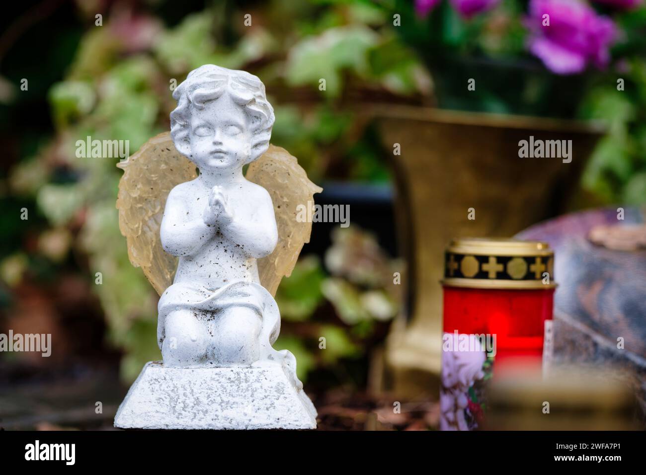 Eine kleine weiße Engelsfigur kniet betend auf einem Grab vor einer Grabkerze und einem Grabstein sowie Blumen im verschwommenen Hintergrund Stockfoto