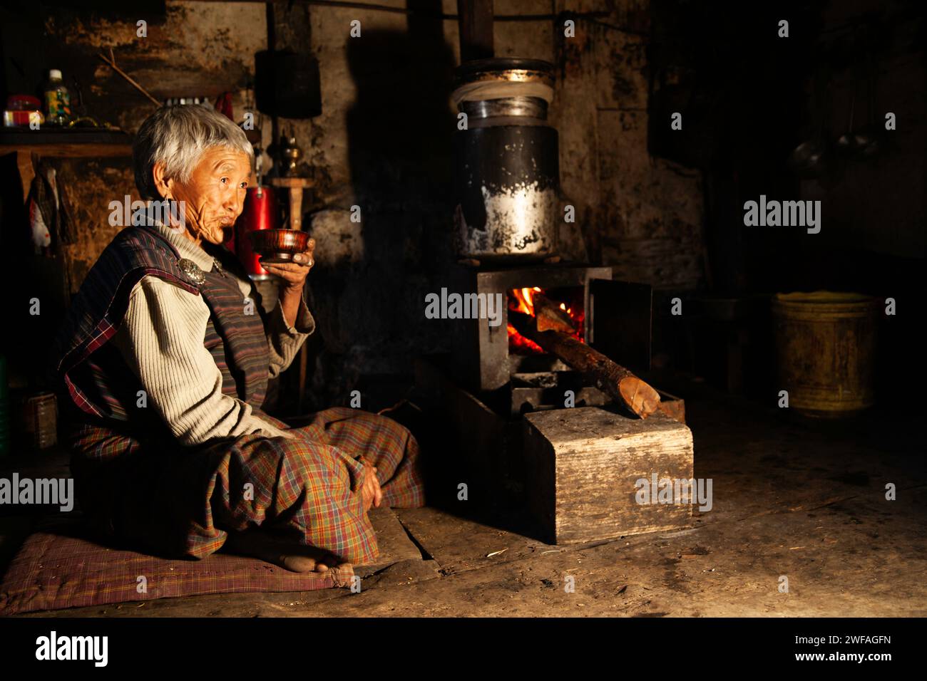 Eine ältere Bhutanese in ihrer rustikalen Küche trinkt frisch zubereiteten, heißen Yak Butter Tee im Dorf Ogyen Choling, Bumthang Valley, Bhutan, Asien Stockfoto