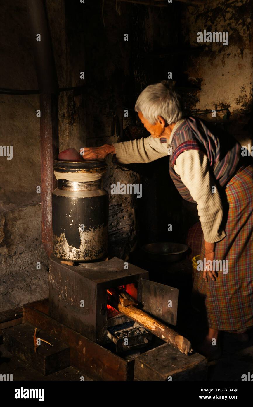 Eine ältere bhutanische Frau in ihrer rustikalen Küche gießt Wasser in einen großen Behälter über ihrem Feuer, um heißen Yakbutter-Tee in Ogyen Choling-Teufel zuzubereiten Stockfoto
