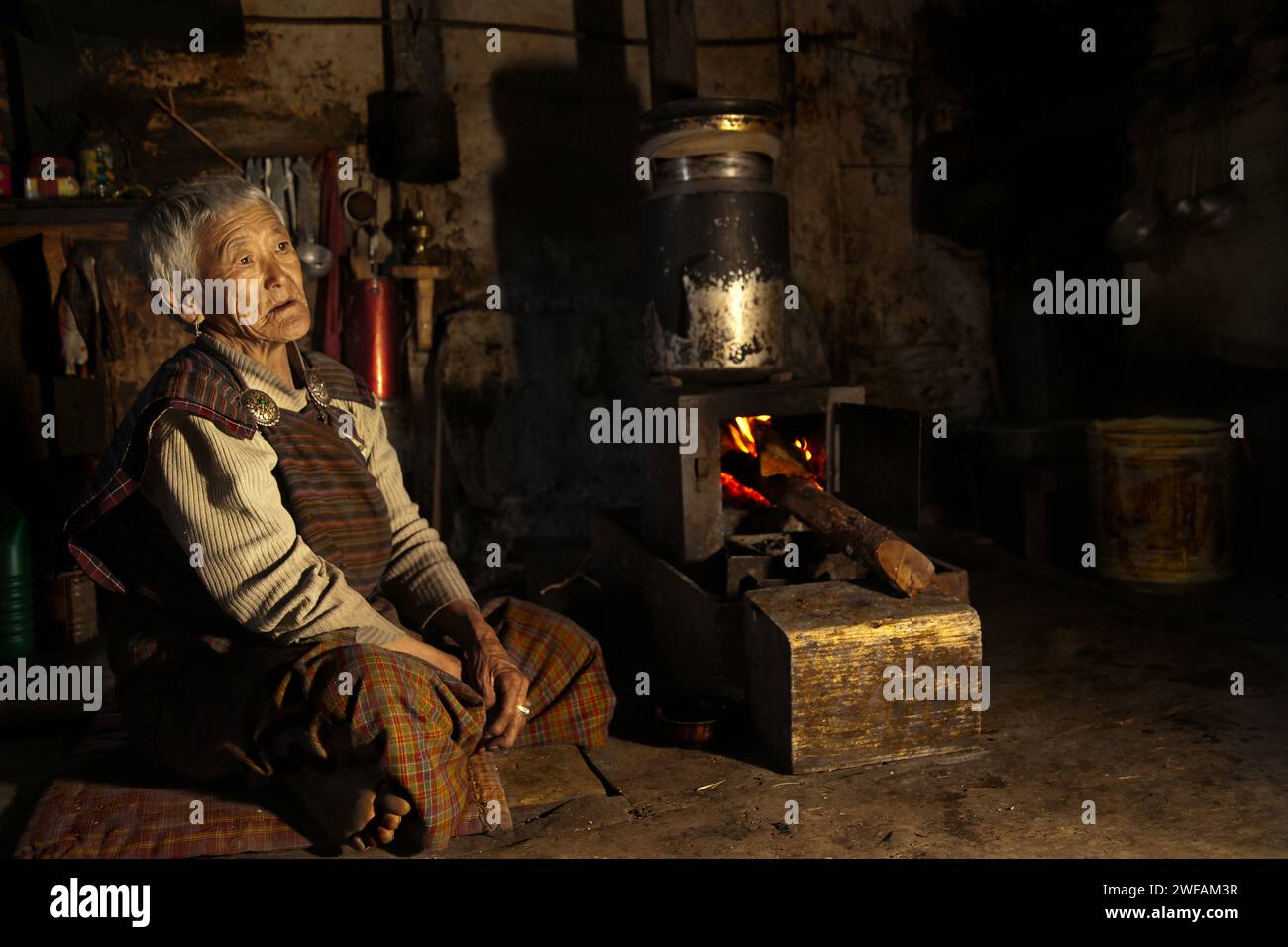 Eine ältere bhutanische Frau in ihrer rustikalen Küche wartet darauf, dass ihr heißer Yak Butter Tee über einem Feuer in Ogyen Choling Dorf, Bumthang, aufgeheizt wird Stockfoto