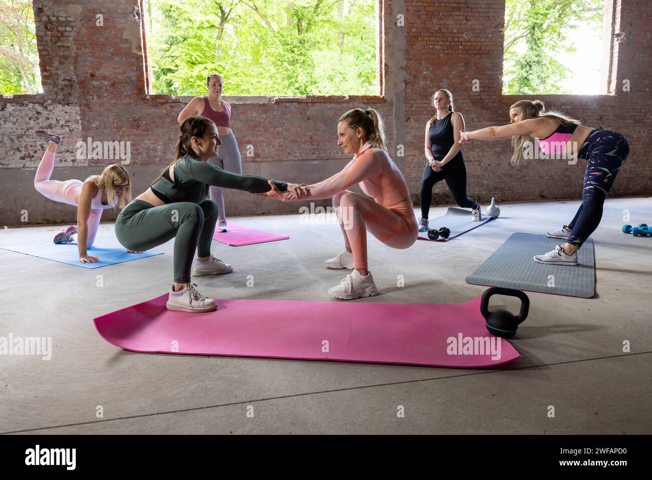 Das Foto zeigt eine Gruppe von Fitnessbegeisterten während eines vielseitigen Workouts in einem geräumigen, urbanen Fitnessstudio. Zwei Frauen stehen an vorderster Front und führen eine Partnerhocke mit gefalteten Händen durch, was Teamarbeit und gegenseitige Unterstützung veranschaulicht. Die umliegenden Teilnehmer werden an verschiedenen Übungen teilgenommen, die einen individuellen Ansatz innerhalb des gemeinschaftlichen Setting zeigen. Der rustikale Charme der freiliegenden Ziegelsteine und die weitläufige Gestaltung des Fitnessstudios unterstreichen die rohe, fokussierte Energie des Fitnessregimes der Gruppe. Teamwork im Training: Partner Squats und Solo-Bemühungen. Hochwertige Fotos Stockfoto